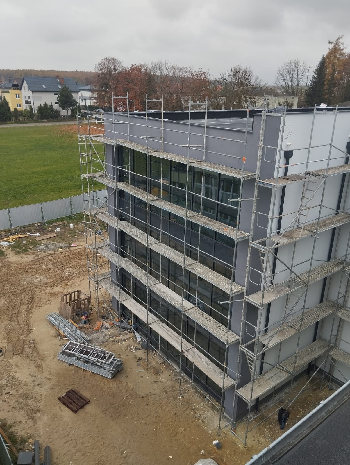 Budynek biurowy w trakcie prac wykończeniowych elewacji, otoczony rusztowaniami, z widocznym terenem budowy i elementami składowanych materiałów.