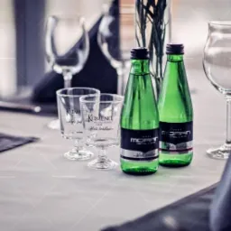 Ustawiony stół z butelkami wody mineralnej w zielonych butelkach, szklankami z logo, kieliszkami i ciemnymi serwetkami, przygotowany do obsługi gości.