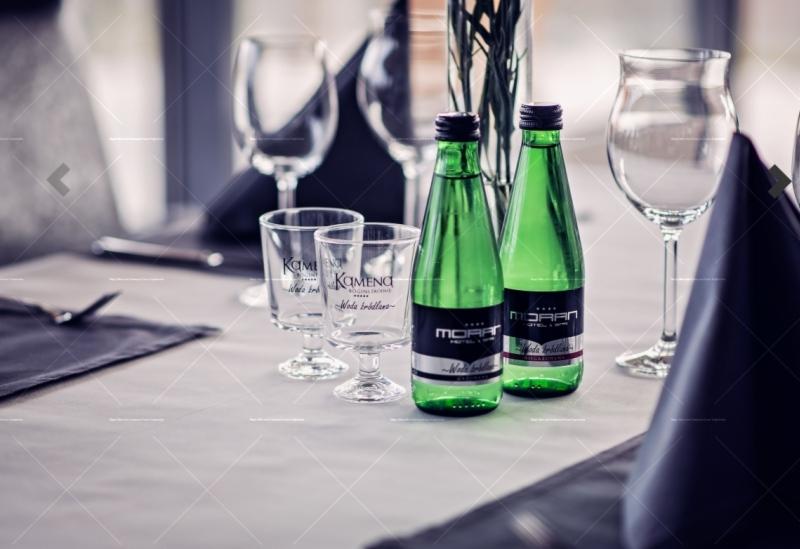 Ustawiony stół z butelkami wody mineralnej w zielonych butelkach, szklankami z logo, kieliszkami i ciemnymi serwetkami, przygotowany do obsługi gości.