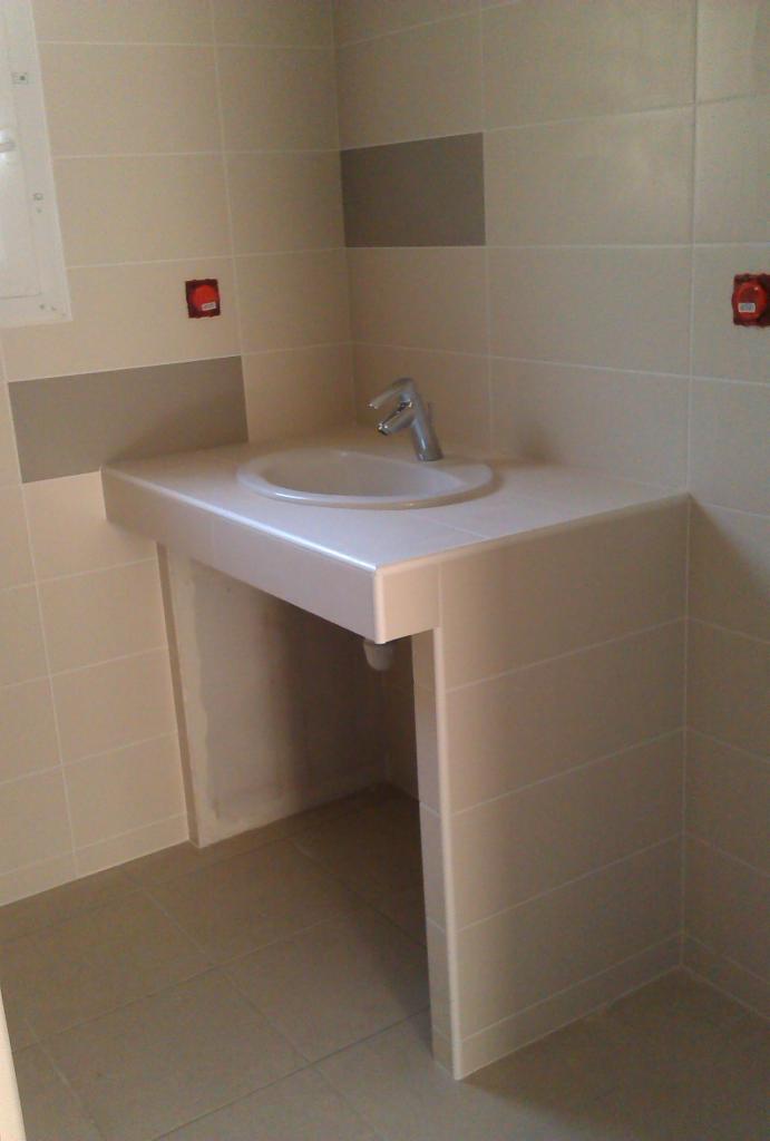 Umywalka ceramiczna z chromowaną baterią, obudowana płytkami w odcieniach beżu i szarości, zamontowana na ścianie wyłożonej kafelkami, z widocznymi puszkami instalacyjnymi.