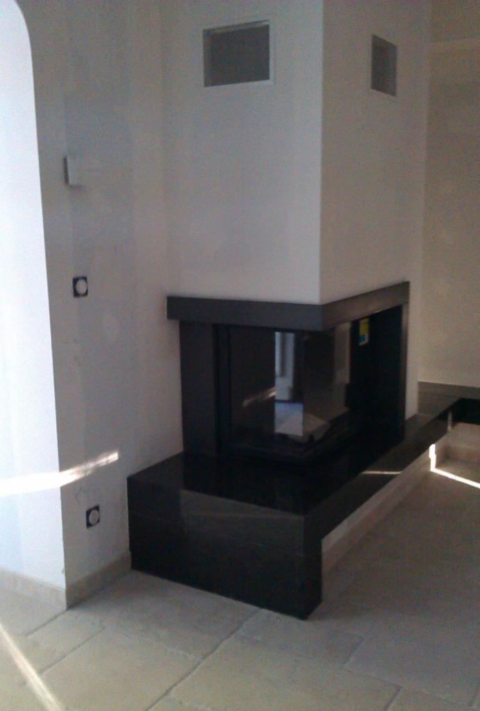 Nowoczesny narożny kominek z czarną obudową, wykończony płytkami podłogowymi w jasnym odcieniu, widoczne gniazdka elektryczne na ścianie.