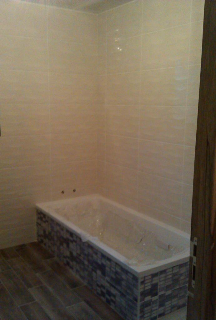 Wykończona łazienka z wanną obudowaną mozaiką, ściany wyłożone jasnymi, beżowymi płytkami, podłoga imitująca drewno, wanna zabezpieczona folią ochronną.