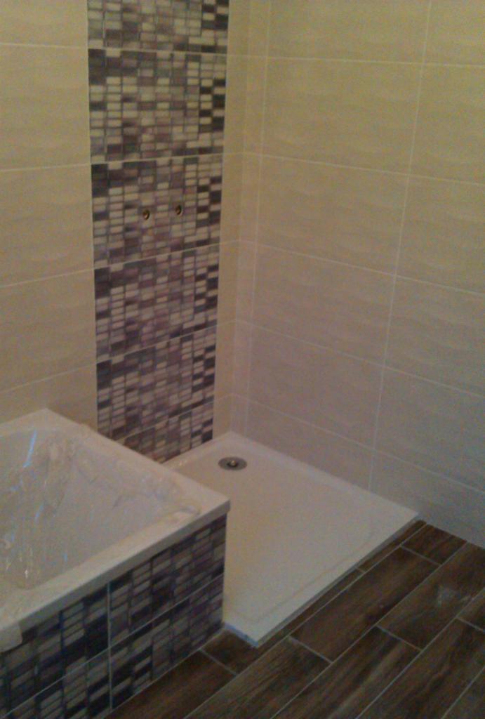 Wykończona łazienka z wanną zabezpieczoną folią, kabiną prysznicową z odpływem i mozaiką na ścianie oraz drewnopodobną podłogą z płytek.