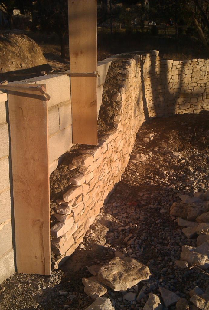 Budowa muru oporowego z kamienia łamanego, widoczne betonowe bloczki i drewniane szalunki, teren budowy z gruzem i ziemią.