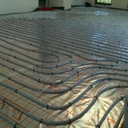 Szare rury ogrzewania podłogowego ułożone na folii z czerwoną kratką w surowym wnętrzu, widok z góry na dużą powierzchnię.