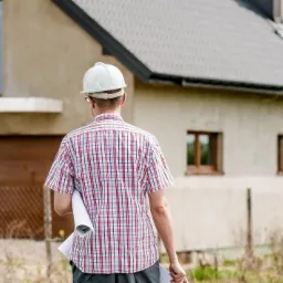 Mężczyzna w kasku budowlanym, z planami w ręku, stoi tyłem do kamery przed domem z oknami i bramą garażową.