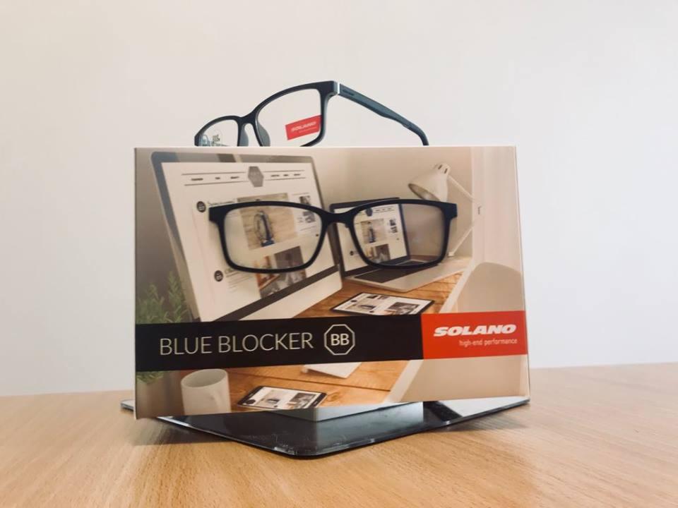Ekspozycja okularów blokujących niebieskie światło marki Solano, prezentowana na kartonowym stojaku z grafiką przedstawiającą biurko z komputerem, tabletem i lampką.