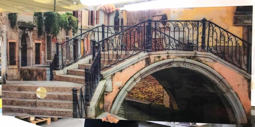 Duży panel z wydrukowanym motywem weneckiego mostu, z widocznymi detalami architektonicznymi i roślinnością, trzymany przez dwie osoby.