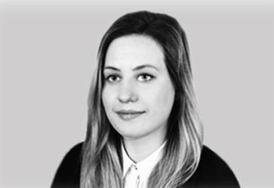 Portret kobiety z długimi włosami w czerni i bieli, uśmiechająca się delikatnie, na jasnoszarym tle. Widok en face, neutralny wyraz twarzy.