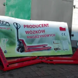 Dwa czerwone wózki paletowe ręczne marki Zakrem ustawione przed banerem reklamowym producenta z logiem firmy i hasłem 'Producent Wózków Magazynowych'.