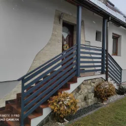 Zewnętrzne schody wejściowe do domu z szarą, poziomą balustradą, wykończone kamieniem i cegłą, z wieńcem świątecznym na drzwiach i śniegiem na roślinach.