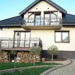 Dwupiętrowy dom z balkonami z kutymi balustradami, widok na ułożone drewno pod balkonem i brukowany podjazd.