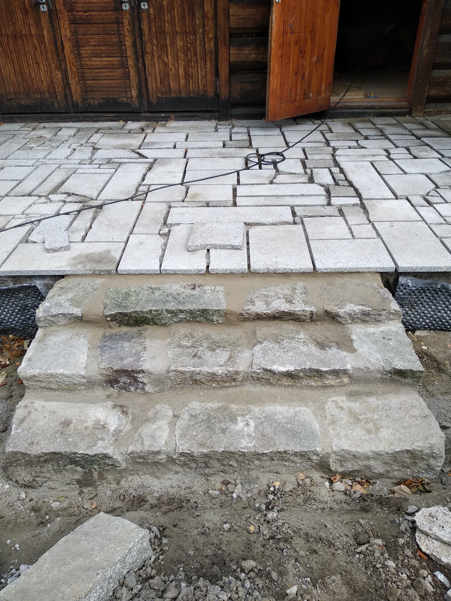 Budowa kamiennych schodów wejściowych z kostki brukowej, widoczne etapy prac ziemnych i układania kostki, z drewnianym budynkiem w tle.