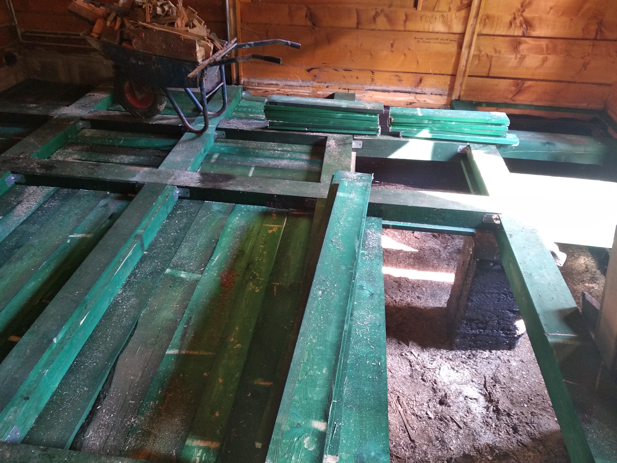 Drewniany szkielet podłogi pomalowany na zielono, częściowo ułożony, z taczką wypełnioną drewnem w tle, widoczne niezagospodarowane podłoże i drewniane ściany budynku.