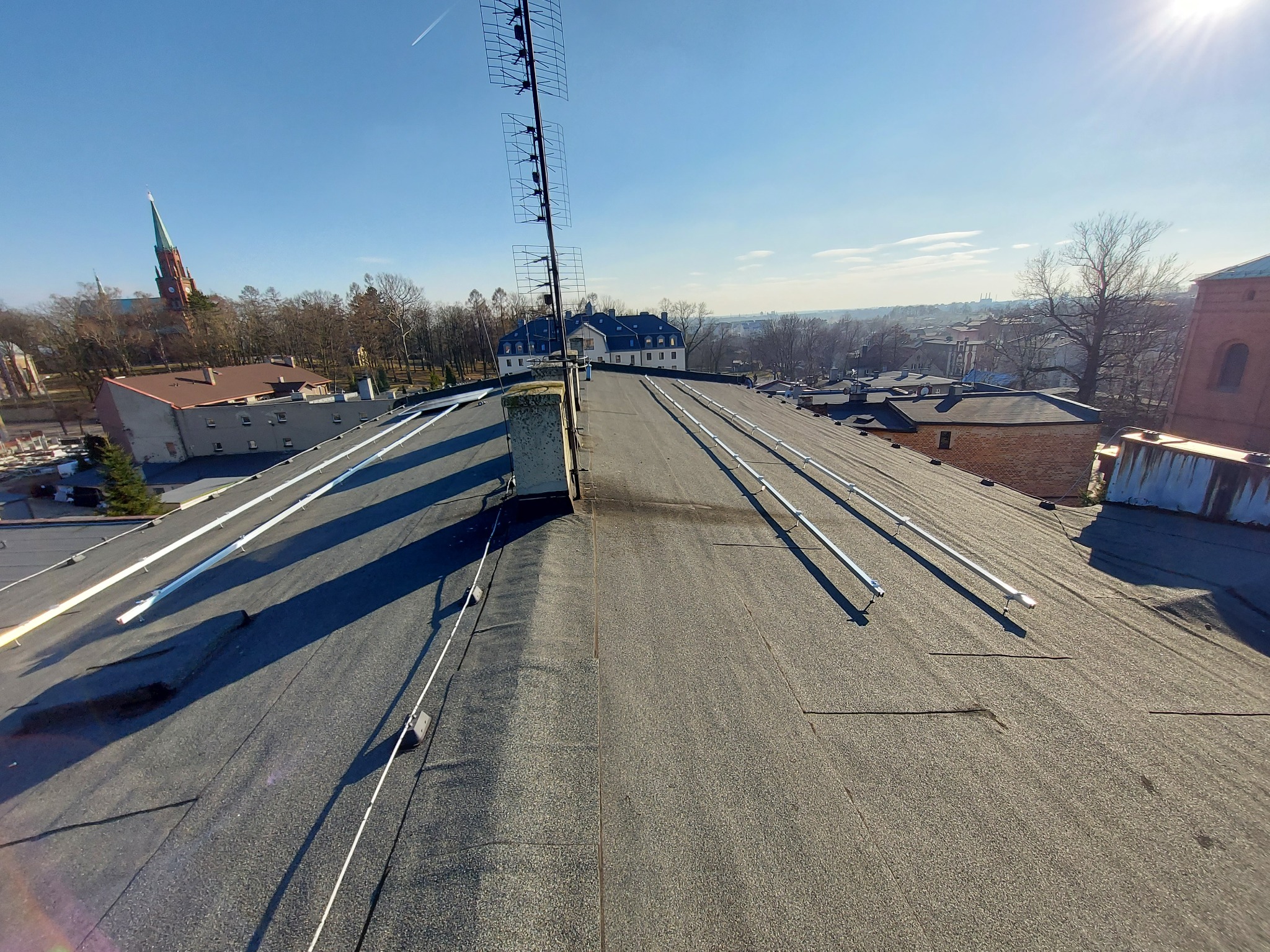 Widok z góry na płaski dach pokryty papą z zamontowanymi elementami instalacji odgromowej i anteną telewizyjną, w tle zabudowania miejskie i kościół