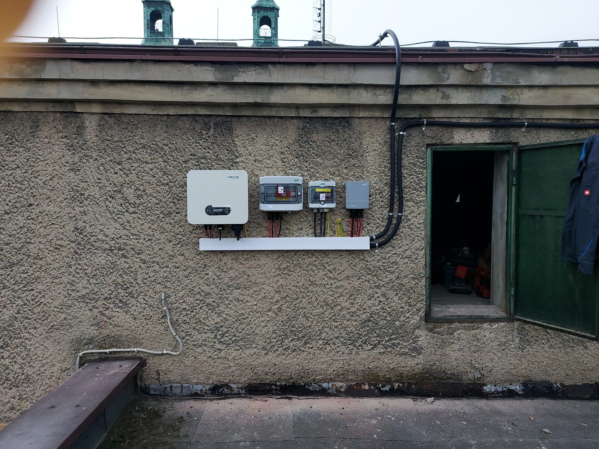 Instalacja paneli słonecznych na dachu budynku z widocznym falownikiem, rozdzielnicami i okablowaniem, na tle zielonych wieżyczek.