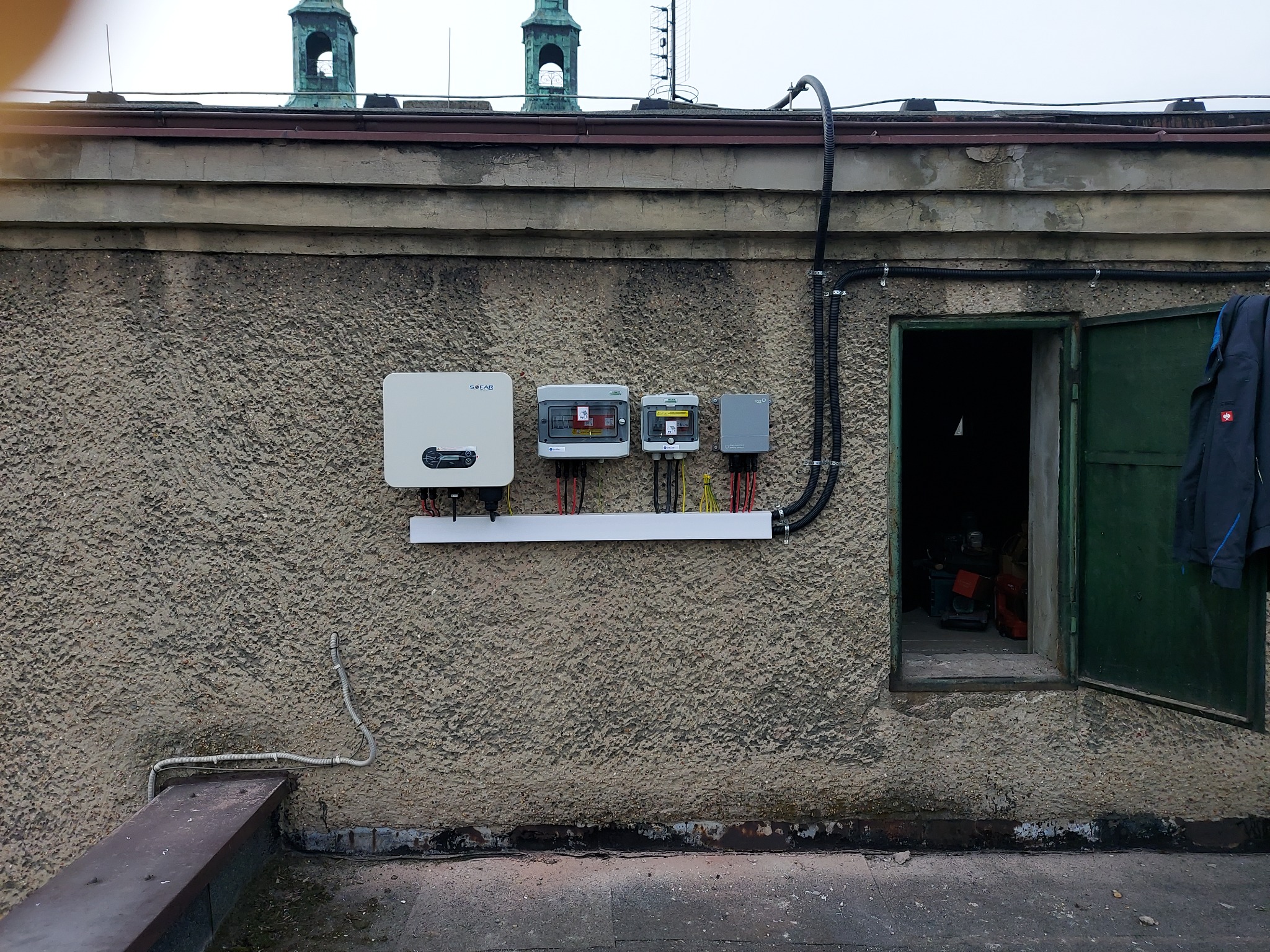 Instalacja paneli słonecznych na dachu budynku z widocznym inwerterem i skrzynkami zabezpieczeń, kable prowadzone w peszlach. W tle widoczne wieżyczki kościelne.