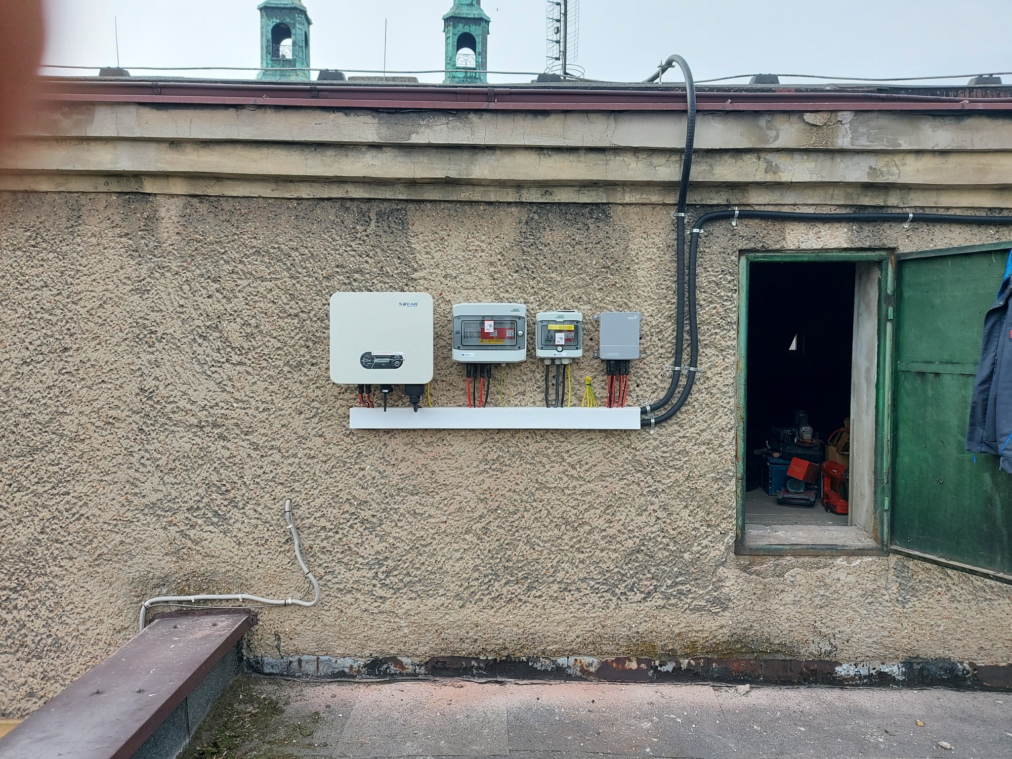 Instalacja fotowoltaiczna na dachu budynku z widocznym inwerterem Sofar i rozdzielnicami, kable w peszlu, w tle wieża kościoła.