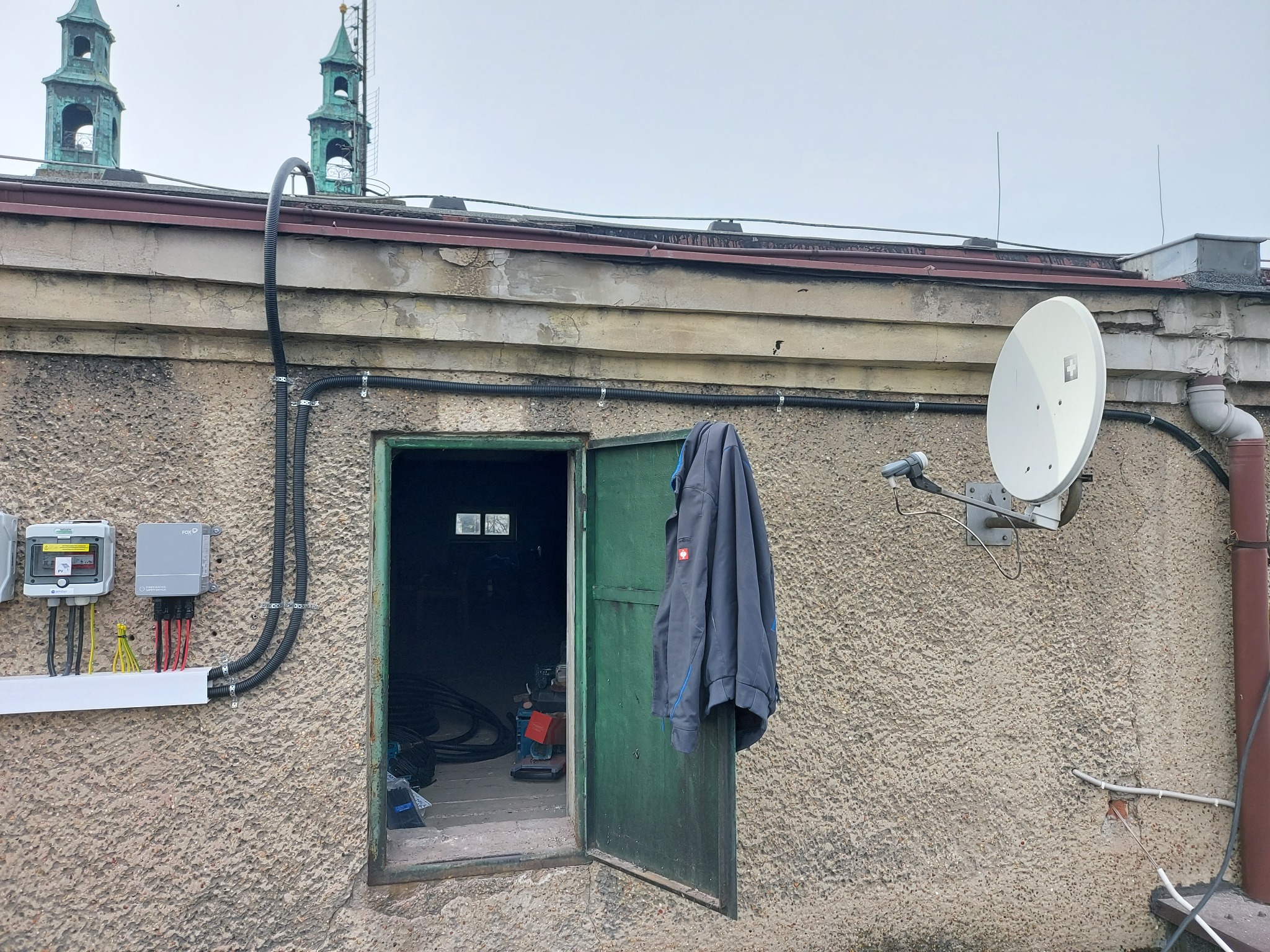 Elewacja budynku z widocznymi elementami: skrzynki elektryczne, kable w peszlach, otwarte drzwi do pomieszczenia z narzędziami, antena satelitarna, zwisająca bluza robocza Engelbert Strauss, w tle...