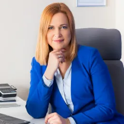 Portret kobiety w niebieskiej marynarce siedzącej przy biurku z laptopem, kalkulatorem i notesem, w tle dyplom w ramce.