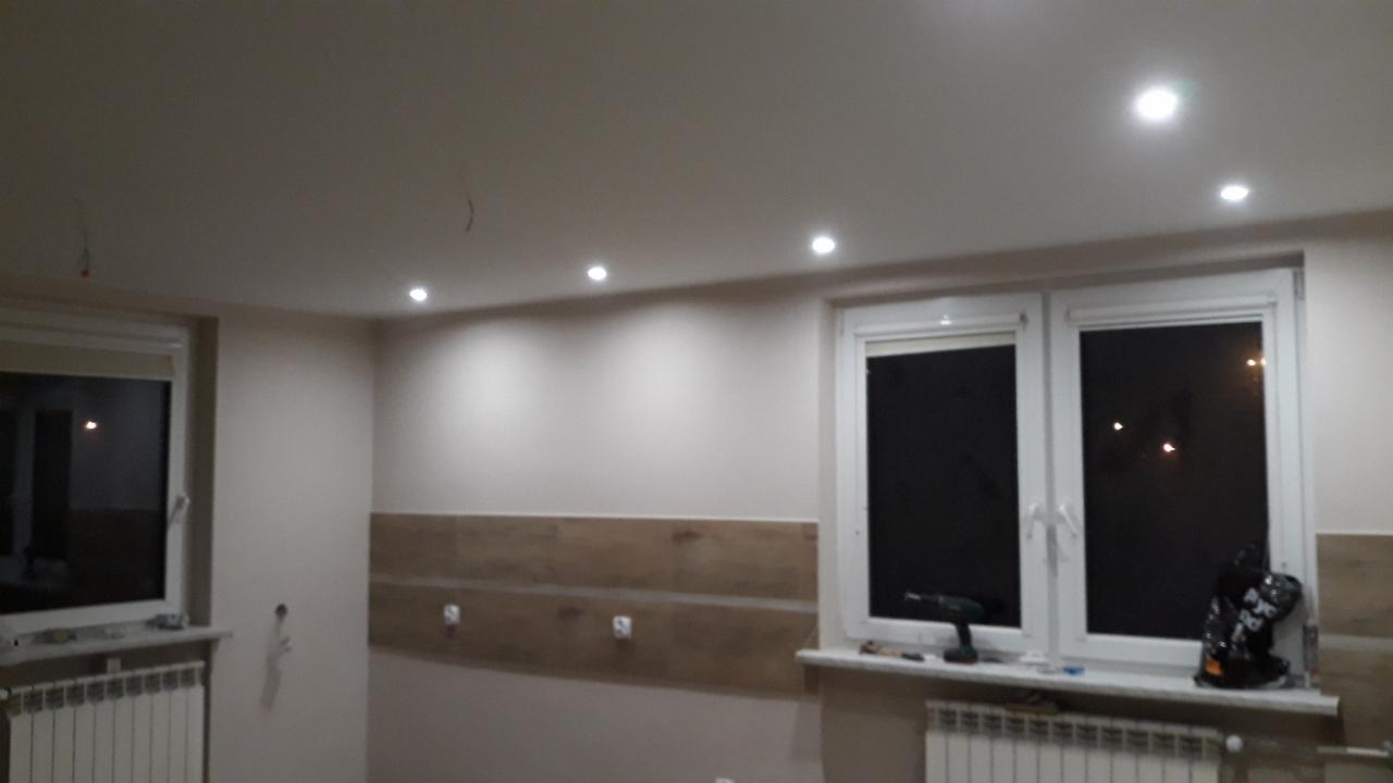 Wnętrze po montażu oświetlenia punktowego w suficie podwieszanym, widoczne niedokończone prace wykończeniowe, w tle okno z narzędziem i torbą na parapecie.