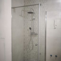 REMKOM SP z O.O. - Kabina prysznicowa z geometrycznymi płytkami w jasnych odcieniach szarości i bieli, widoczne niepodłączone krany i wystający kabel elektryczny.