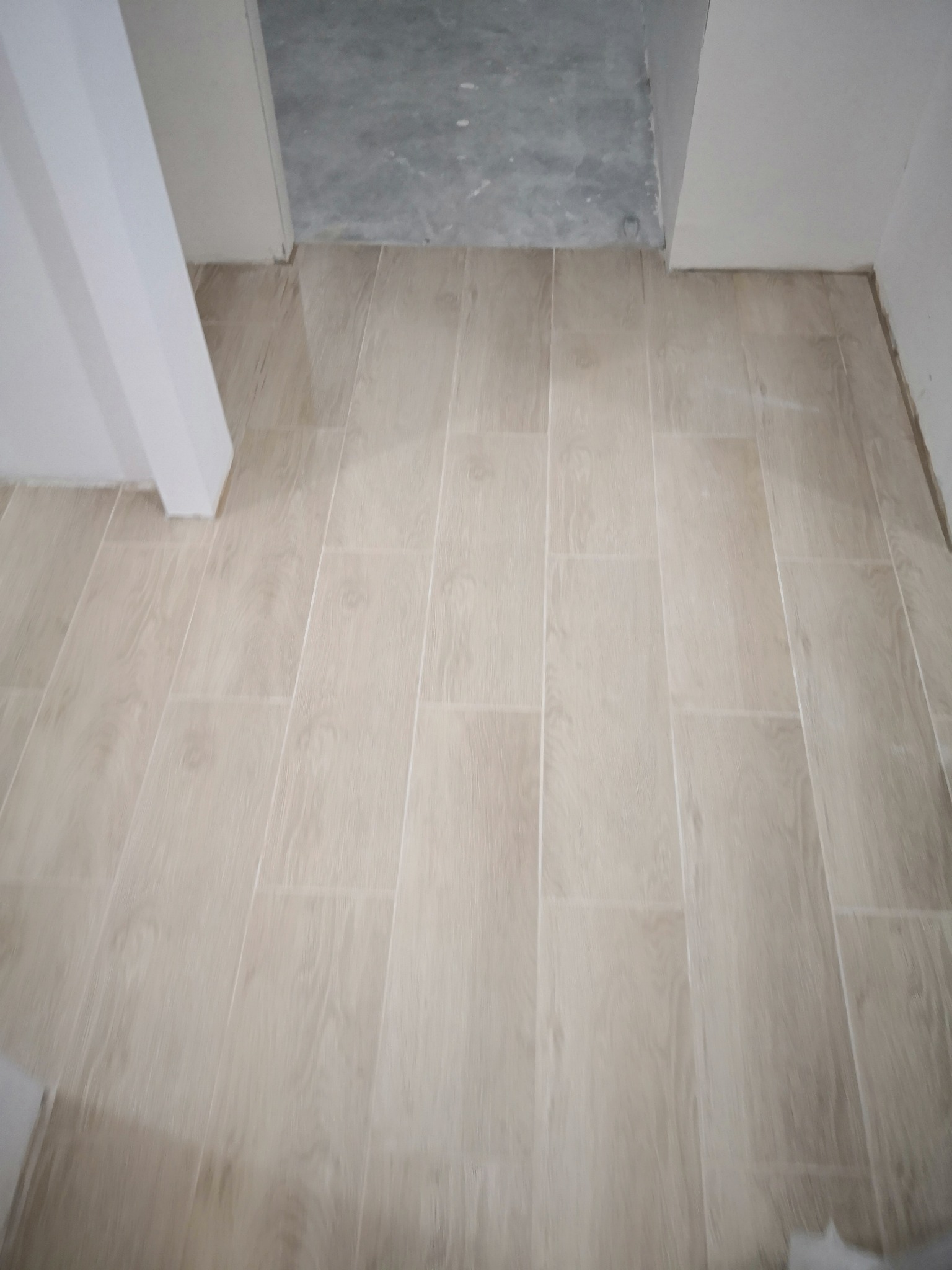 Ułożone jasne płytki podłogowe imitujące drewno, widoczne fugi, w tle surowy beton na podłodze w przejściu.