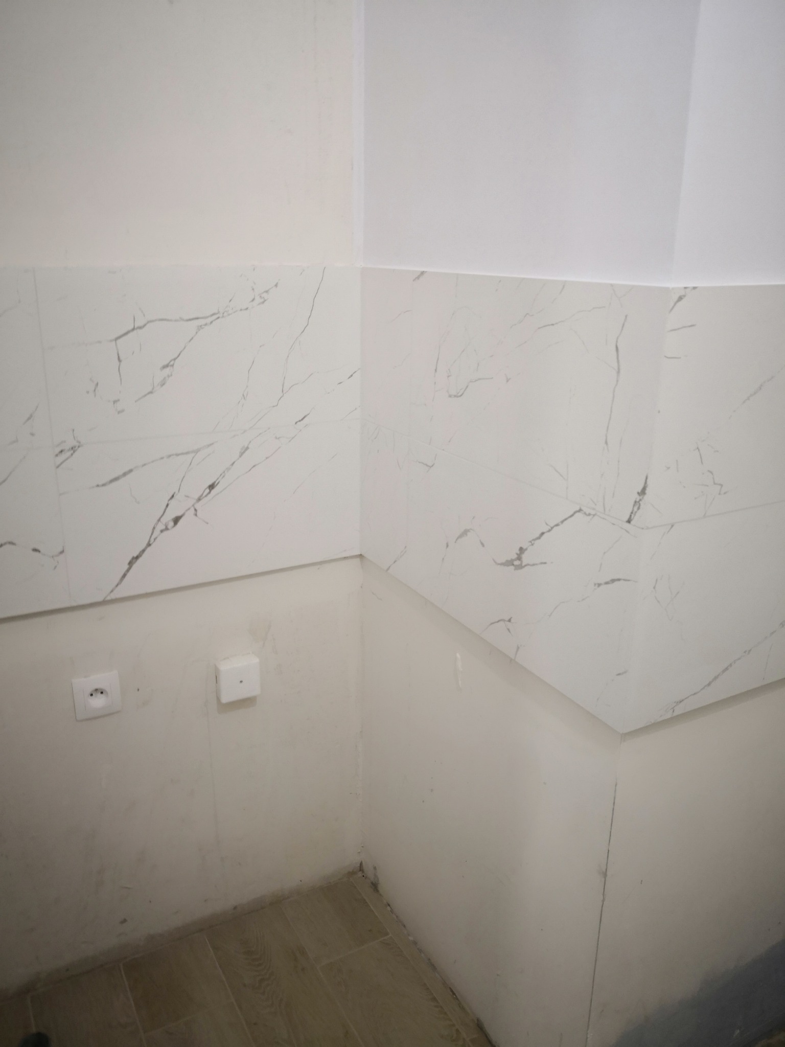 Fragment narożnika łazienki w trakcie wykańczania: częściowo położone płytki imitujące marmur na ścianie, widoczne gniazdka elektryczne, surowy stan ściany poniżej płytek i fragment podłogi.