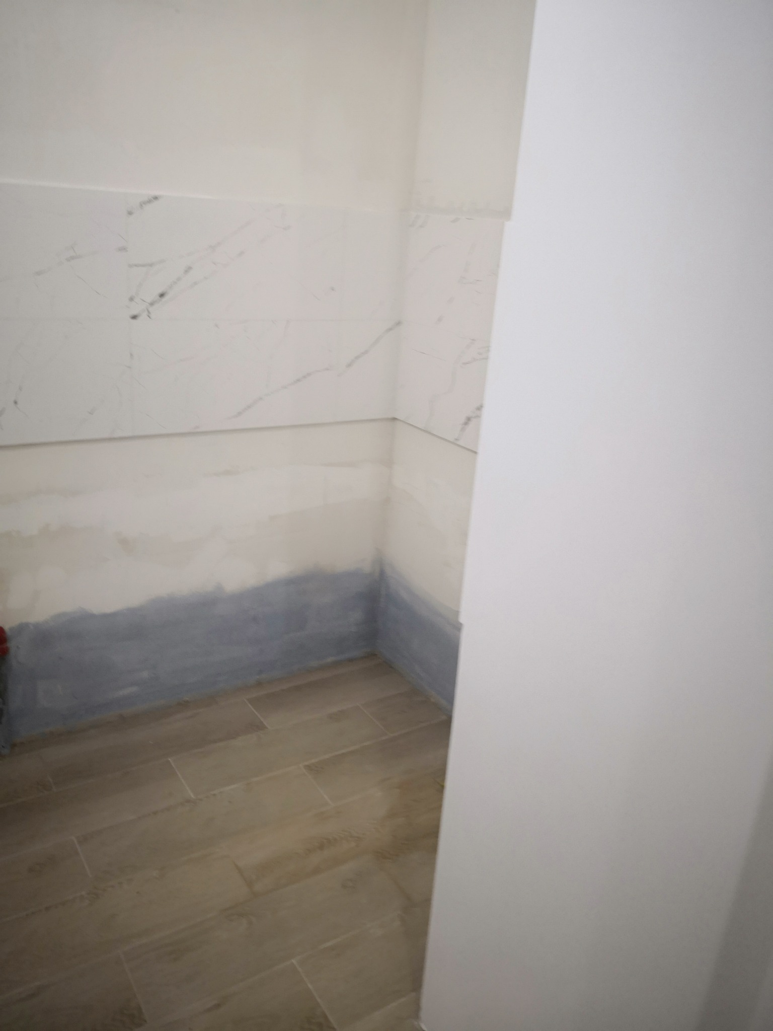 Ściana łazienki w trakcie przygotowania do wykończenia: widoczne płytki imitujące marmur w górnej części, dolna część ściany z szarą warstwą izolacyjną, podłoga wyłożona płytkami imitującymi drewno.