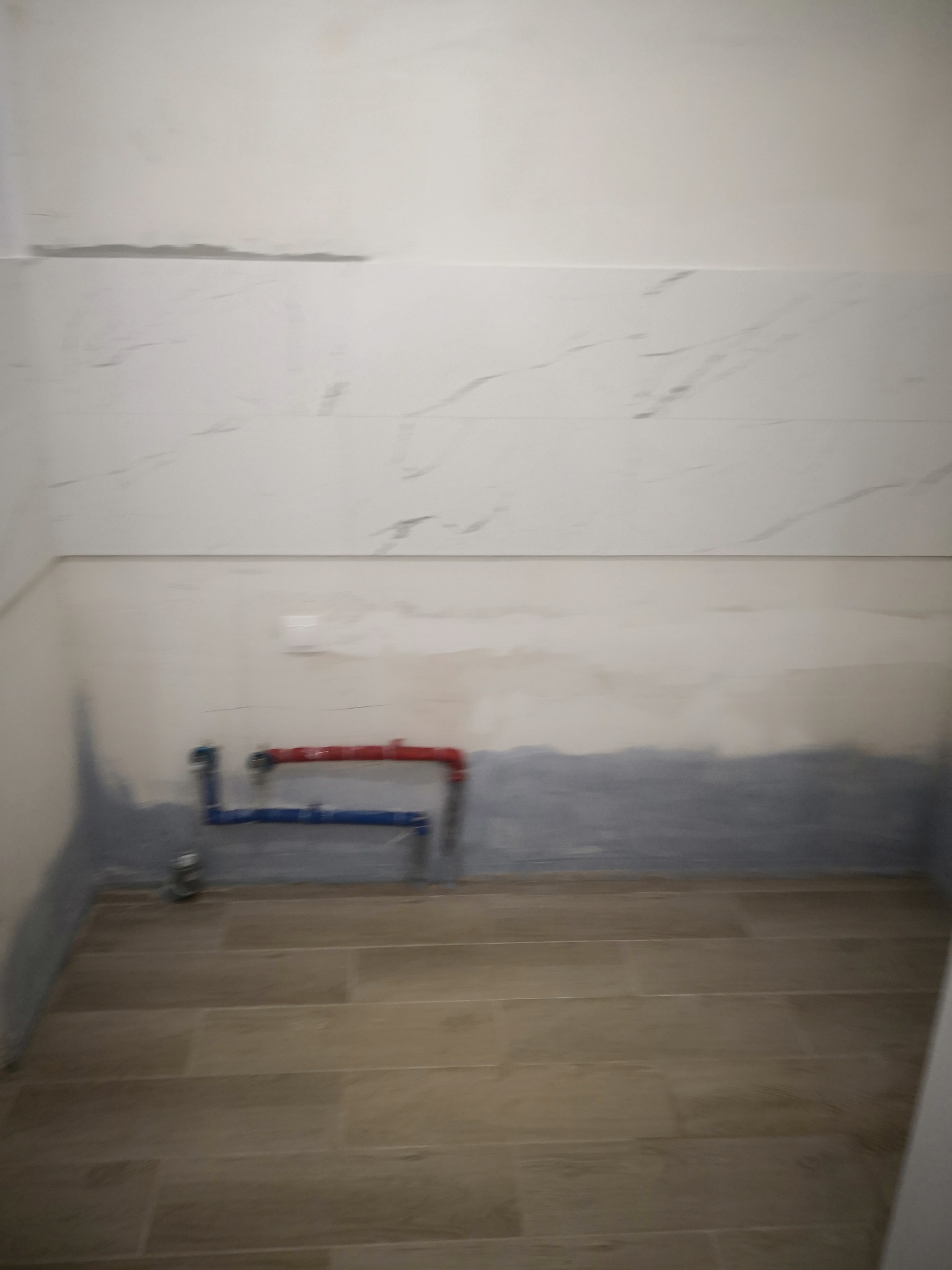 Niedokończona łazienka z położoną podłogą z płytek imitujących drewno, częściowo wykończonymi ścianami z płytek imitujących marmur i widocznymi rurami instalacji wodnej.