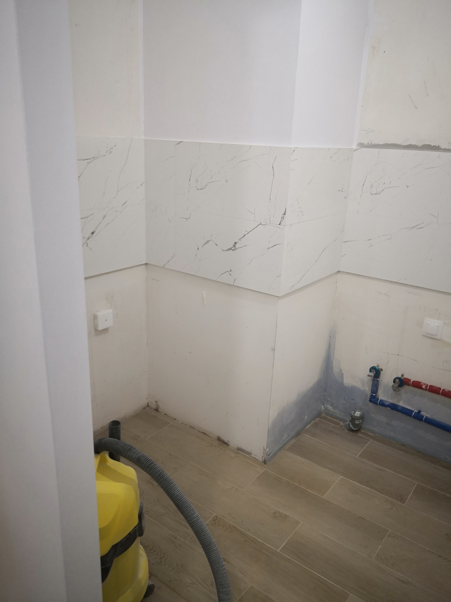 Łazienka w trakcie wykańczania: częściowo położone białe płytki imitujące marmur na ścianach, widoczne rury instalacji wodnej, fragment żółtego odkurzacza przemysłowego i drewnopodobna podłoga...