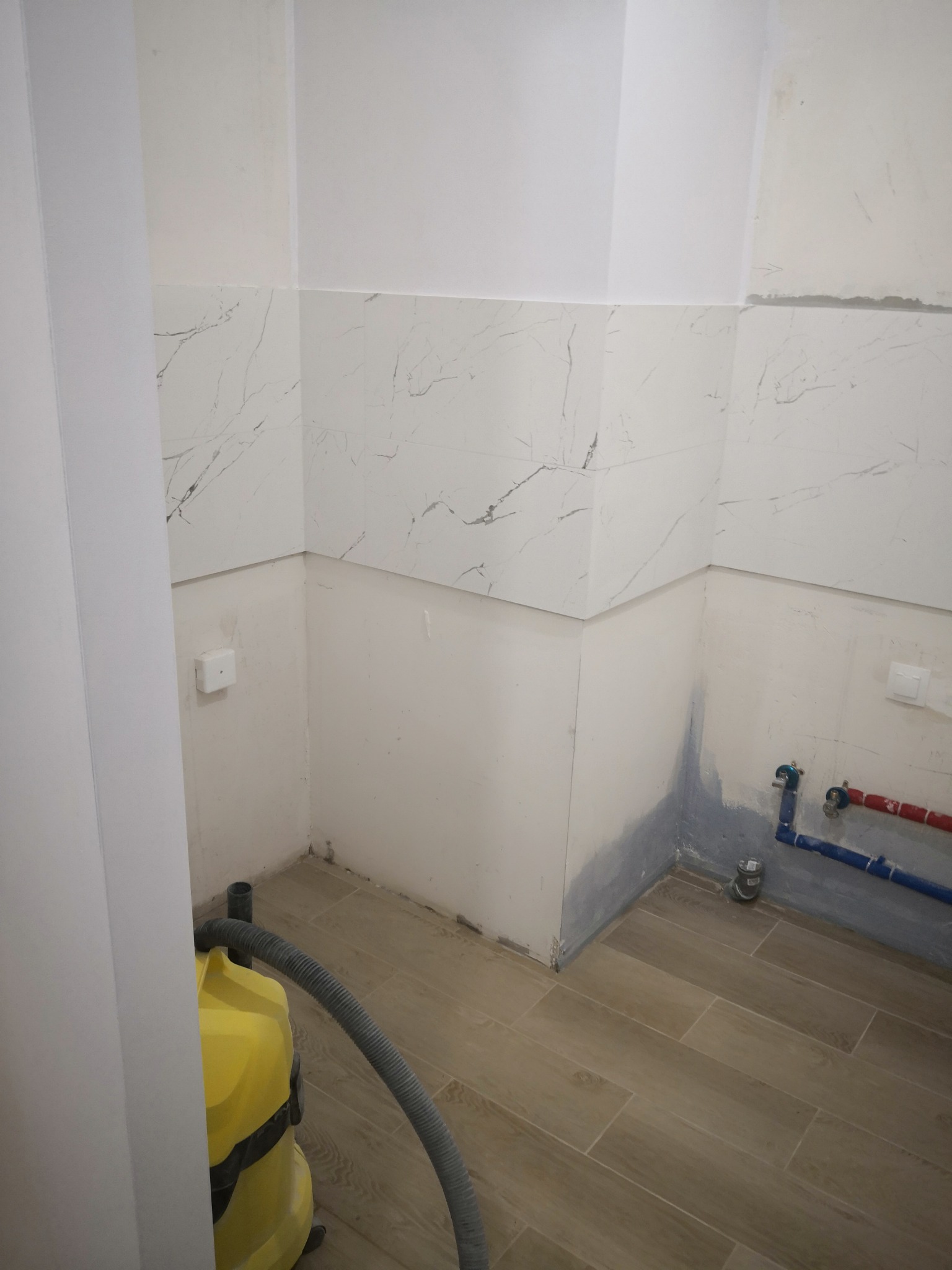 Łazienka w trakcie prac wykończeniowych: częściowo położone płytki imitujące marmur na ścianach, przygotowane ściany do malowania, widoczne rury instalacji wodnej i odkurzacz przemysłowy.