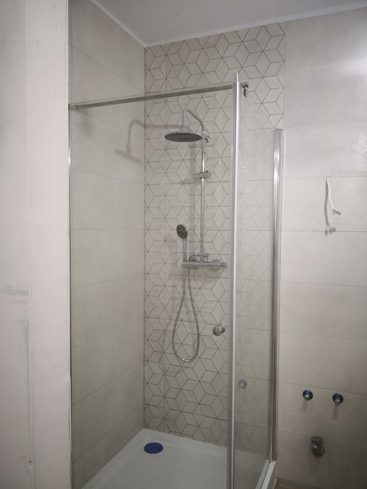 Kabina prysznicowa z geometrycznymi płytkami w jasnych odcieniach szarości i bieli, widoczne niepodłączone krany i wystający kabel elektryczny.