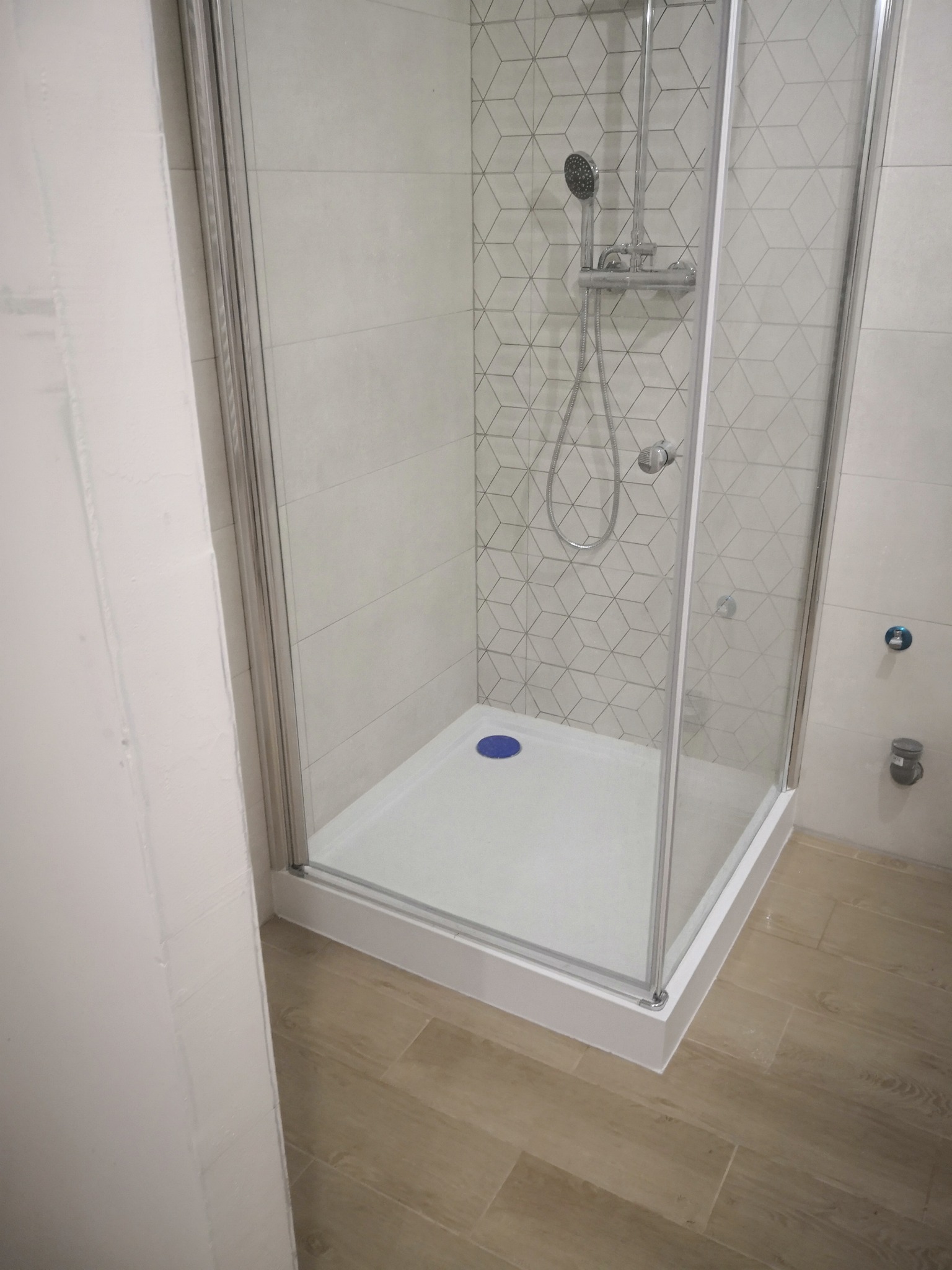 Nowoczesna kabina prysznicowa z geometrycznym wzorem na płytkach, zamontowana w narożniku łazienki, widoczny odpływ z niebieskim akcentem