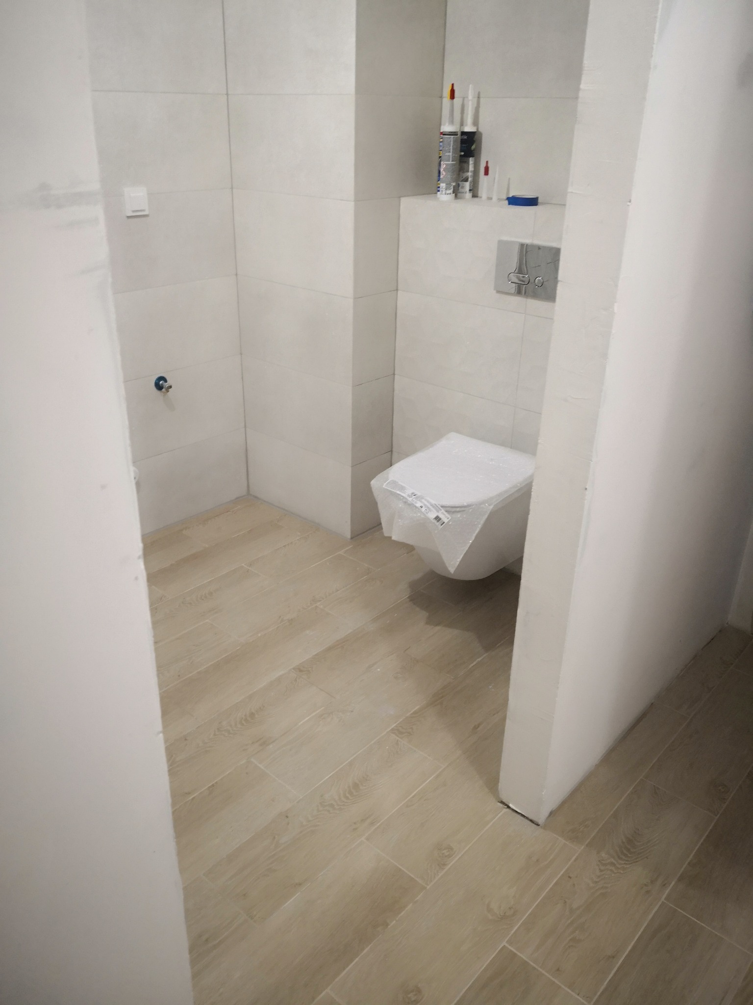 Nowa, częściowo wykończona łazienka z beżowymi płytkami imitującymi drewno na podłodze; toaleta w folii ochronnej, na półce tuby silikonu i przycisk spłuczki.