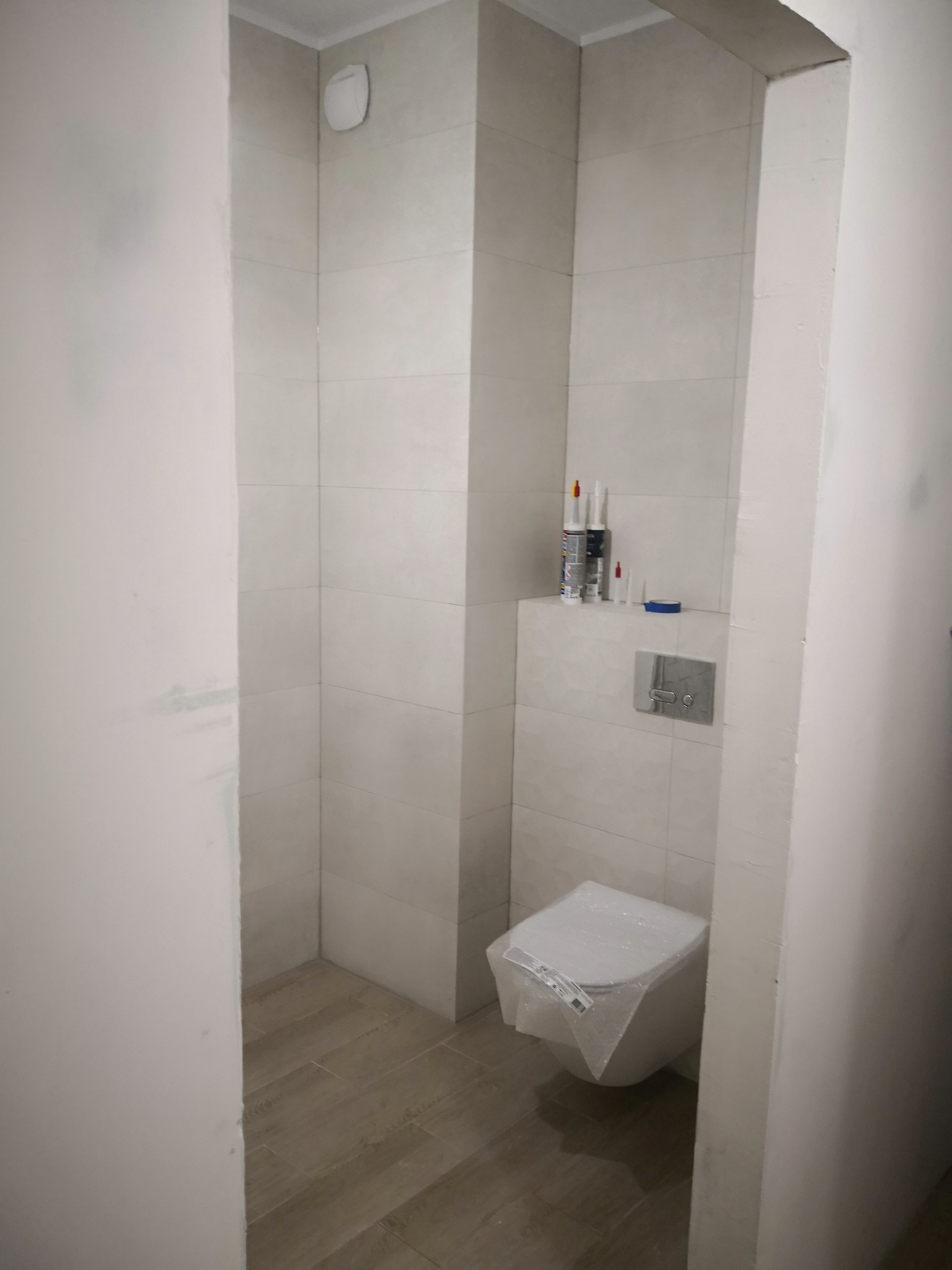 Wnętrze łazienki w trakcie remontu: częściowo wykończone płytki ścienne w odcieniach bieli, brązowa podłoga, podwieszana toaleta jeszcze w folii ochronnej i materiały budowlane na półce.