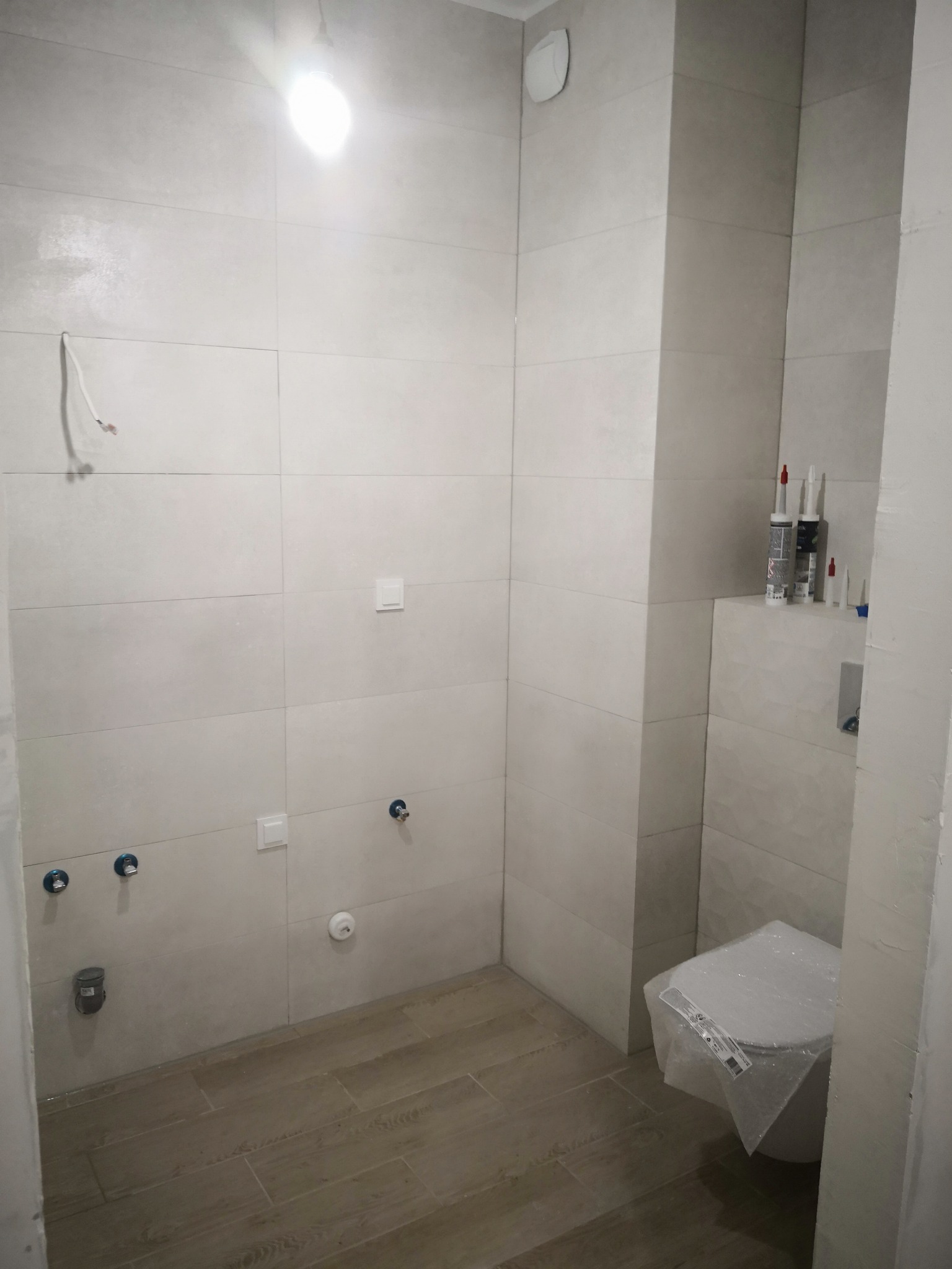 Łazienka w trakcie wykończenia: ułożone płytki ścienne i podłogowe w odcieniach szarości, widoczne przyłącza hydrauliczne, zabezpieczona folią toaleta, silikony budowlane na półce, surowy kabel...