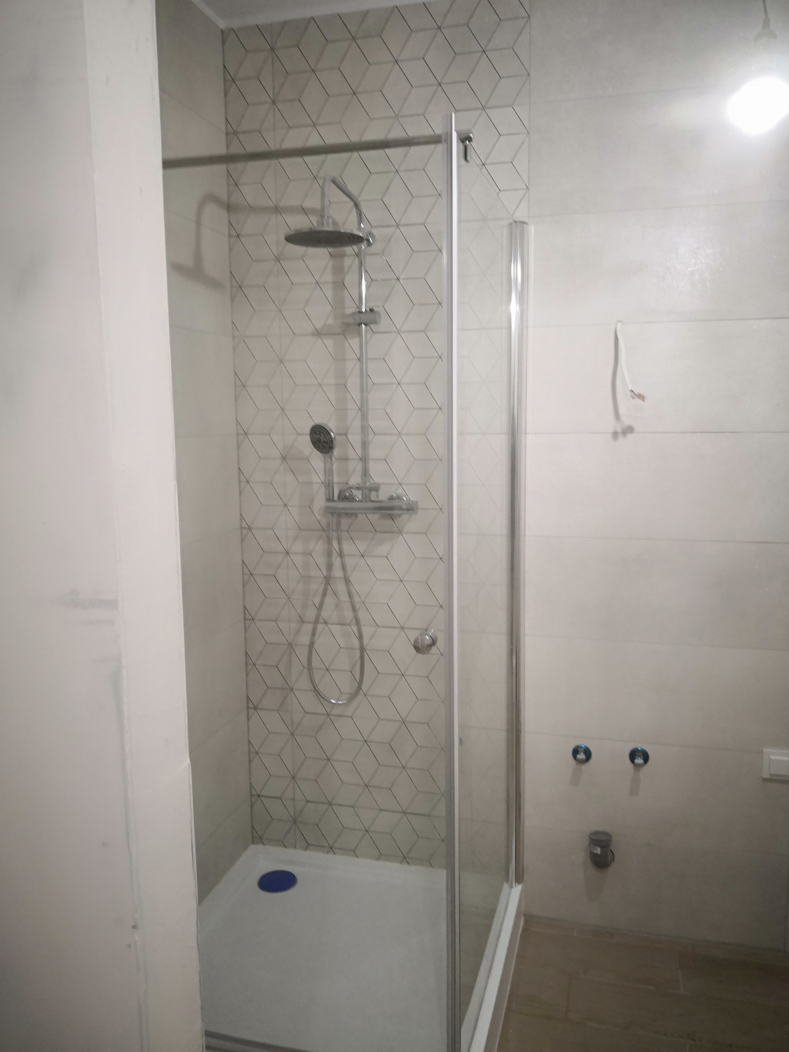 Kabina prysznicowa z deszczownicą i baterią termostatyczną na tle ściany wyłożonej geometrycznymi płytkami; widoczne niedokończone podłączenia wodne na ścianie obok.