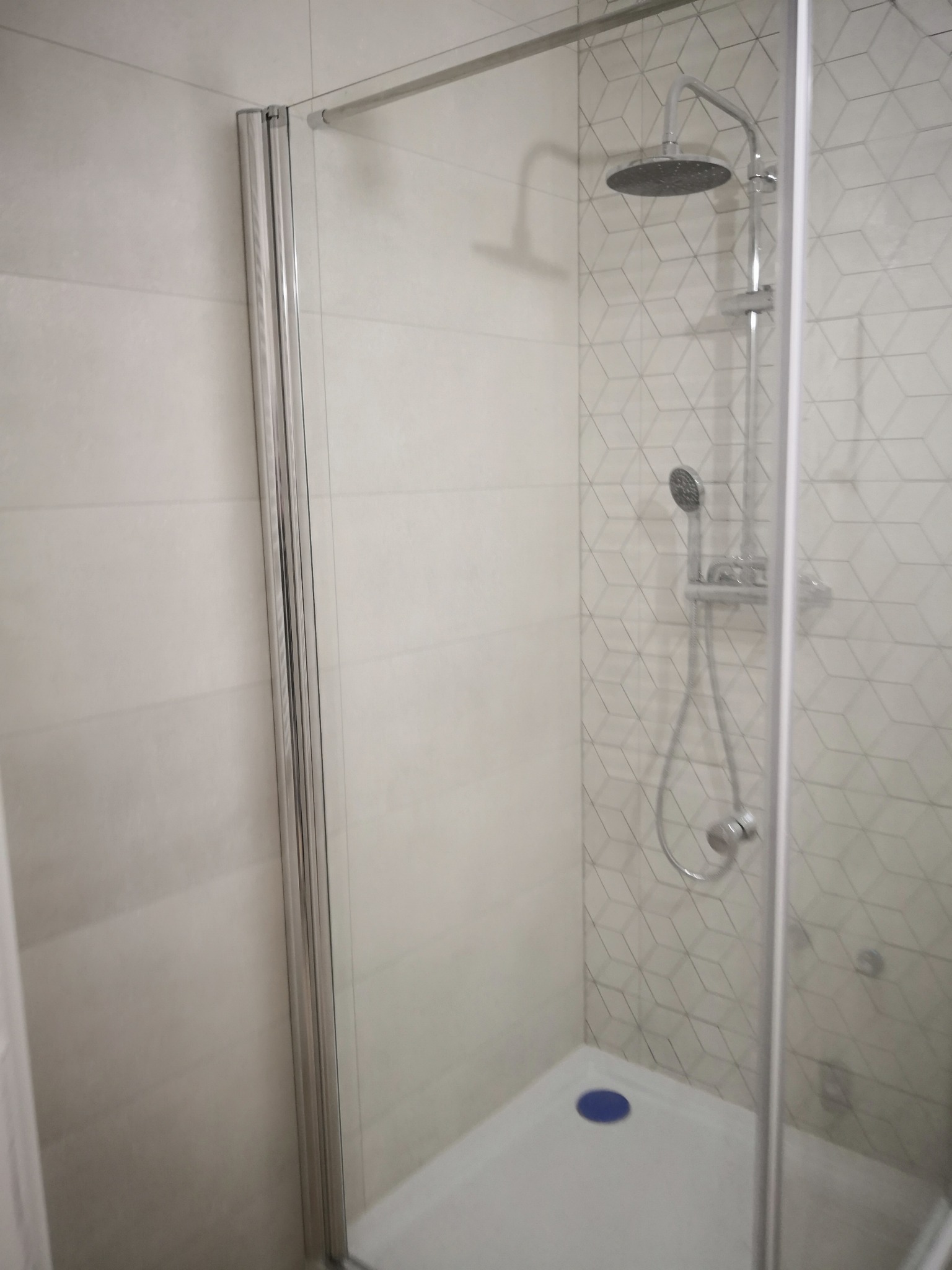 Kabina prysznicowa z deszczownicą i słuchawką, białe płytki z geometrycznym wzorem, widoczny niebieski odpływ w brodziku.