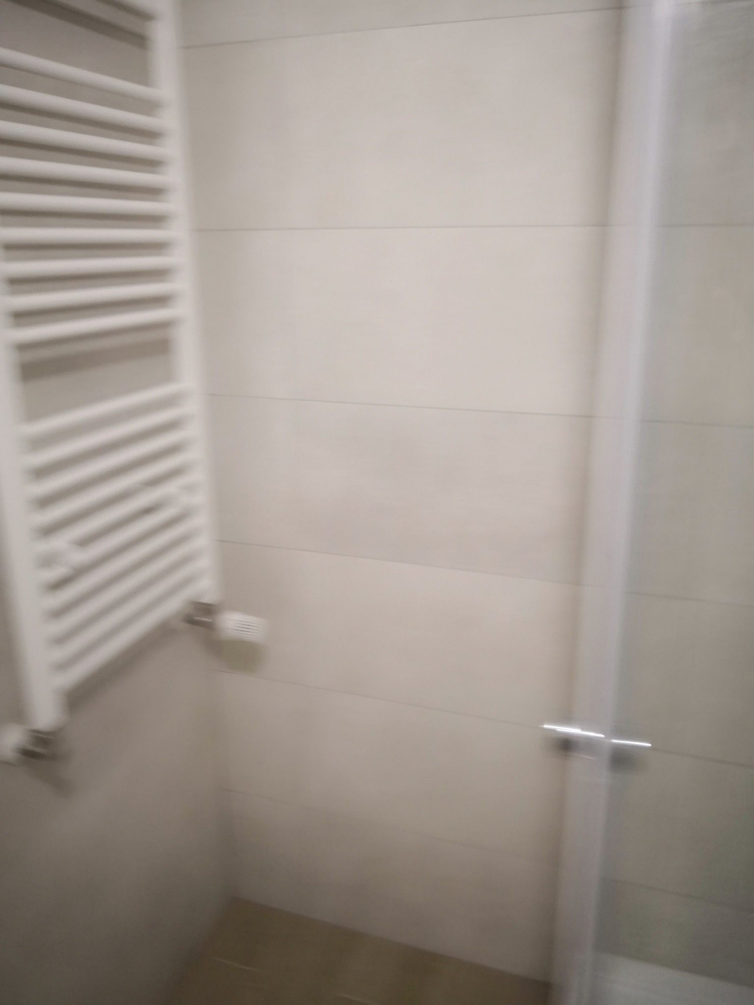 Biały grzejnik drabinkowy na ścianie z beżowych płytek w łazience, obok kabina prysznicowa z aluminiową ramą, widok z dołu.