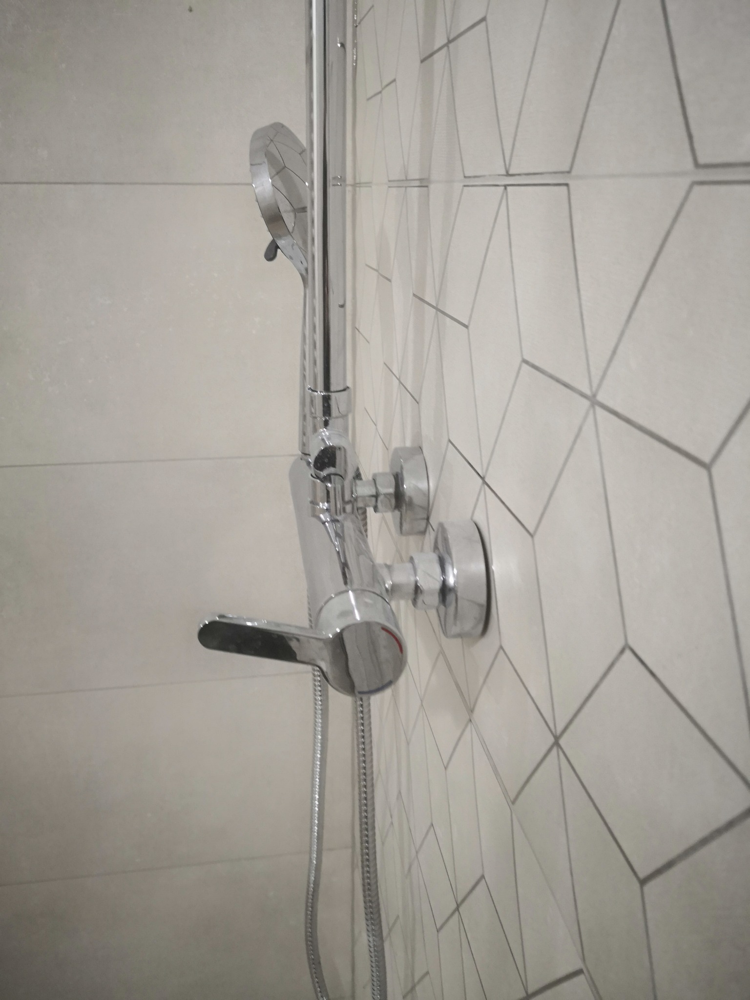 Bateria prysznicowa z deszczownicą i wężem natryskowym zamontowana na ścianie wyłożonej jasnymi płytkami z geometrycznym wzorem.