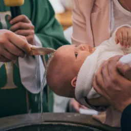 Ksiądz polewa wodą głowę niemowlęcia trzymanego przez rodzic&oacute;w podczas ceremonii chrztu, w tle zielona szata liturgiczna.