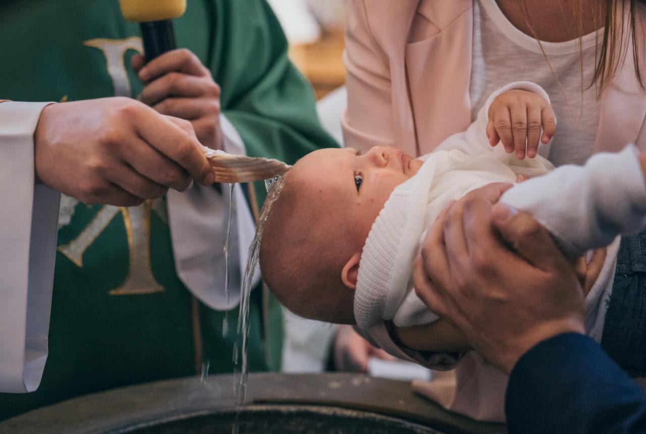 Ksiądz polewa wodą głowę niemowlęcia trzymanego przez rodziców podczas ceremonii chrztu, w tle zielona szata liturgiczna.