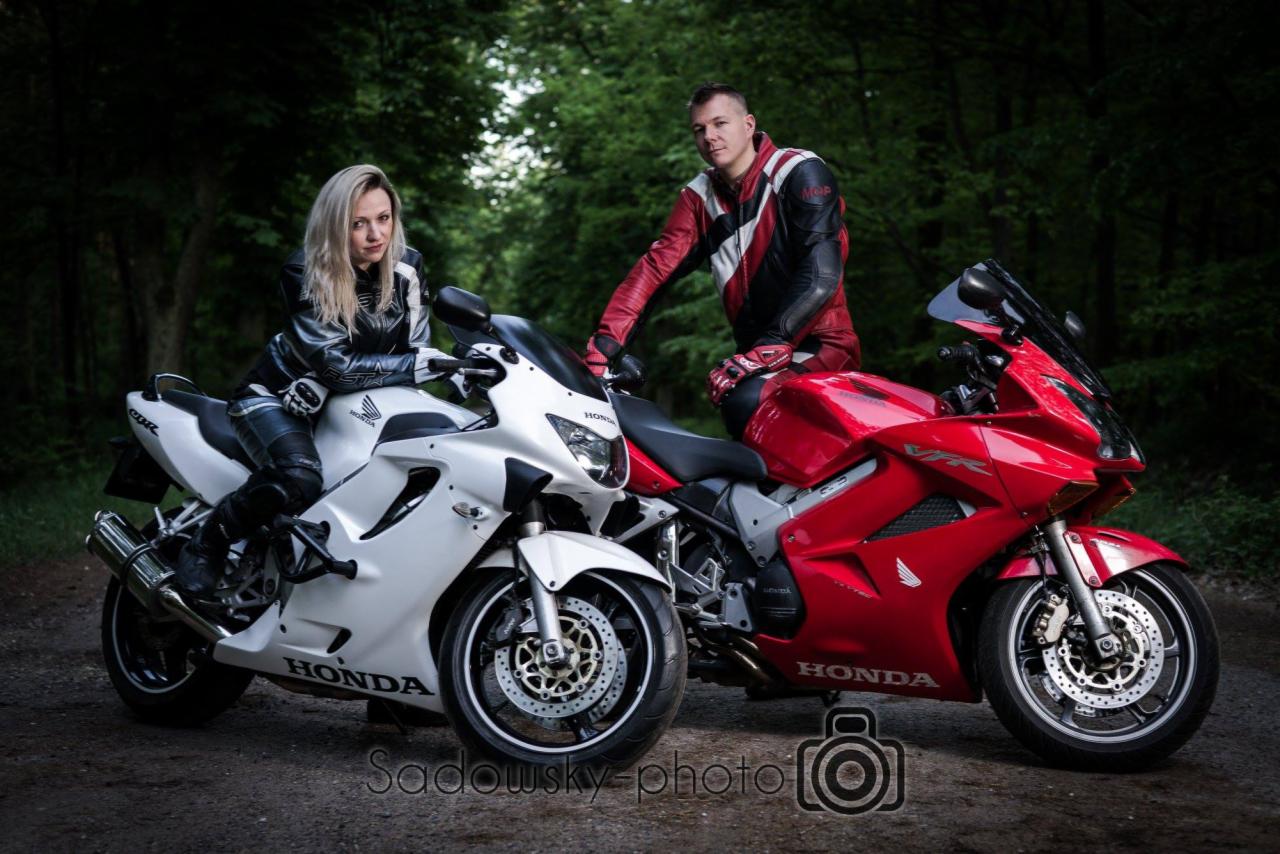 Para w skórzanych strojach motocyklowych pozuje z dwoma motocyklami Honda: biały i czerwony, na leśnej drodze.