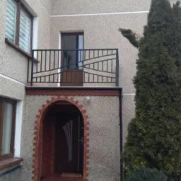 Elewacja budynku z balkonem zabezpieczonym czarną, metalową balustradą o geometrycznym wzorze, nad wejściem z ceglanym łukiem.