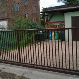 Przesuwna brama wjazdowa z metalowymi prętami, brązowa rama, dekoracyjny motyw p&oacute;łkola, tło z budynkiem z czerwonej cegły i garażem.