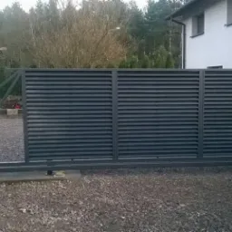 Przesuwna brama wjazdowa z poziomych, metalowych profili, szara, widoczna szyna i mechanizm jezdny, na żwirowym podłożu, w tle dom i drzewa.