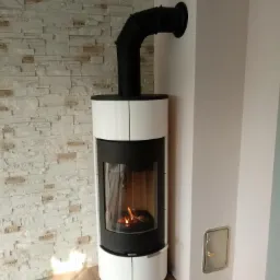 Nowoczesny piec wolnostojący z białymi panelami i czarnym wykończeniem, podłączony do komina, z widocznym płomieniem w palenisku, na tle ściany z dekoracyjnej cegły i pomalowanej ściany z małą...