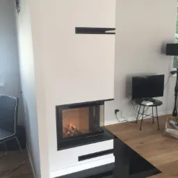 Nowoczesny kominek narożny z czarnymi akcentami, palenisko z płomieniami, na podłodze parkiet, w tle telewizor na stoliku.