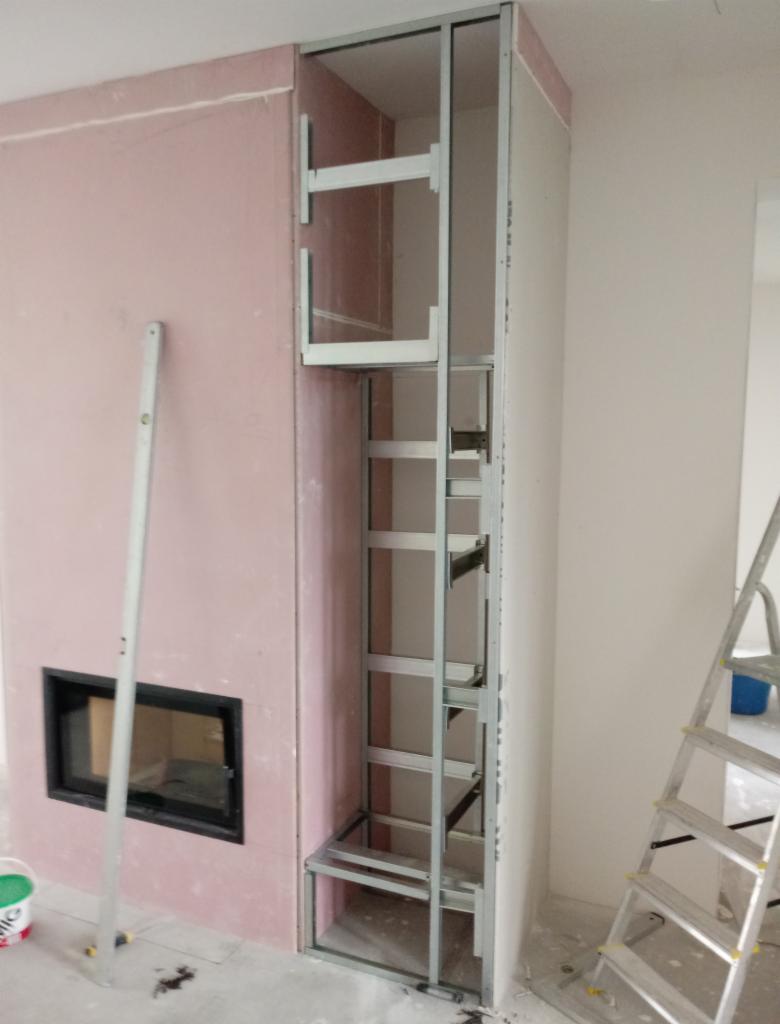 Konstrukcja metalowa obudowy kominka z zamontowanym wkładem, poziomica oparta o ścianę, widoczny etap prac wykończeniowych.